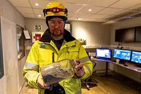 Gruvearbeider i gult verneutstyr holder et glass med malmprøve i et kontrollrom.