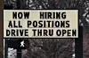 Roadside sign reading Now Hiring All Positions above a walk signal at an intersection.
