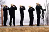 Five people in straw hats use binoculars and a scope to watch birds by a foggy reservoir.