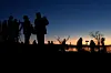 Silhouetted birdwatchers with cameras and tripods at a reservoir at dawn.