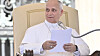 A pope in white vestments sits in an ornate chair and holds papers near a microphone.
