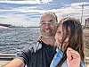 Two people take a selfie by the water with a cloudy sky and distant city skyline behind them.