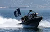 A rigid inflatable boat carrying Turkish Navy commandos speeds across the water with a coastal city behind it.