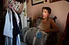 Mujer mayor sentada en una silla de ruedas junto a una bolsa y ropa colgada en una habitación.