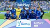 Jugadores de Millonarios posan en el campo junto a un panel de BetPlay.