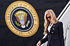 Katie Zacharia in a navy tailored suit and sunglasses descends Air Force One beside the presidential seal.