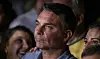 Jair Bolsonaro aparece em primeiro plano, cercado por pessoas, em um ambiente interno.